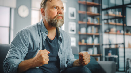 Charismatic Bearded Man Sitting on a Couch Watches Game on TV, Worrying for His Soccer Team. Focused, Joyfully Intense and Authnetic Fan Cheers when Favourite Club Play