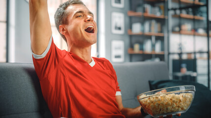 Handsome Young Adult Man Sitting on a Couch Watches Soccer Game on TV, Cheers for his Team to Win...