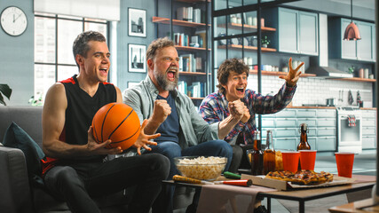 At Home Three Happy Basketball Fans Sitting on a Couch Watch Game on TV, Celebrate Scoring and...