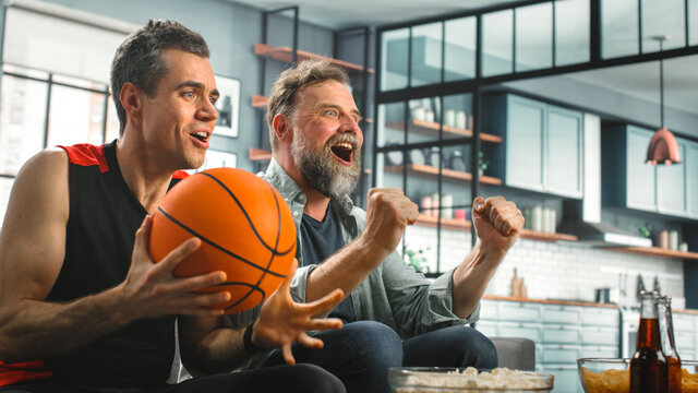 At Home Two Fans Sitting On A Couch Watch Basketball Game On TV, Celebrate Scoring Victory. Thier Team Wins Championship. Friends Watch Favourite Club Play