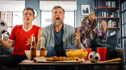 At Home Three Joyful Soccer Fans Sitting on a Couch Watch Game on TV, Celebrate Victory when Sports Team Wins Championship. Group of Friends Cheer When Favourite Football Club Play. Portrait Shot.