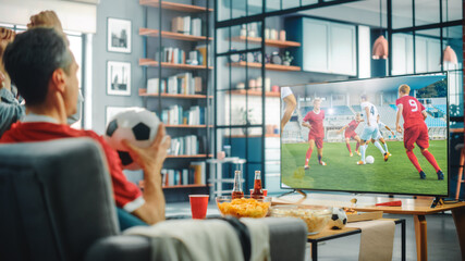 At Home Soccer Fans Sitting on a Couch Watch Football Game on TV, Cheer for Favourtite Sports Team to Win Championship. Screen Shows Professional Football Club Play. Over the Shoulder