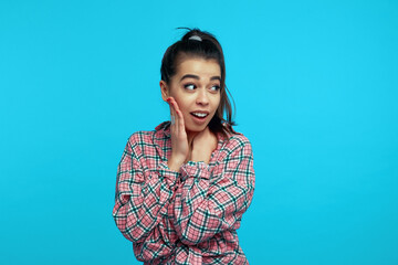 Fototapeta premium Beautiful wondered woman keeps hand on cheek looks surprisingly aside dressed in trendy clothes reacts on shocking news isolated over blue background