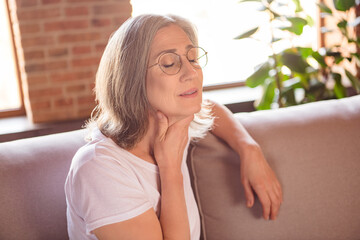 Fototapeta premium Portrait of attractive sick grey-haired woman sitting on divan touching neck feeling bad at home house flat indoors
