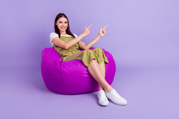 Full body photo of cheerful young happy woman point finger empty space sit bean bag isolated on purple color background