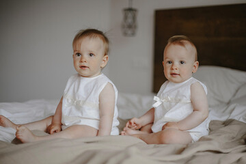 Front view of cute children siblings sitting on white bed and looking straight. Beautiful baby...