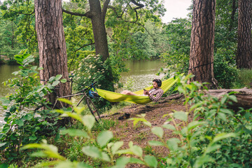 Young caucasian man resting in hammock, listening to music on headphones and using smartphone afterwards on bicycle in forest near the lake. Audio healing. Idyllic place. Travel, camping in nature