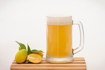 Jarra de cerveza con refresco de limón y limones con hojas, sobre una tabla de madera y fondo blanco.