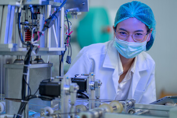 Factory worker woman with gown uniform stand near part of mask producing machine and look at camera with smiling under hygiene mask in her face.