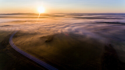 View from above. An asphalt road is located among endless green meadows. Automobile road in the fog against the background of dawn.