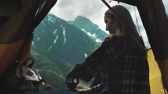 A young girl hiker drinks tea from a thermos in the mountains. Tea drinking outdoors. Woman sitting and drinking tea in yellow tent, camping adventure lifestyle outdoors