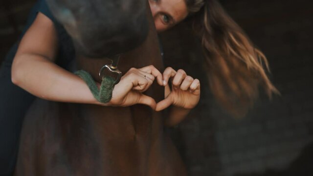 Horsewoman holding her dark bay horse with love and making a heart with her fingers. Ways of expressing love for horses. Girl with arms around her horse standing in the stable. 