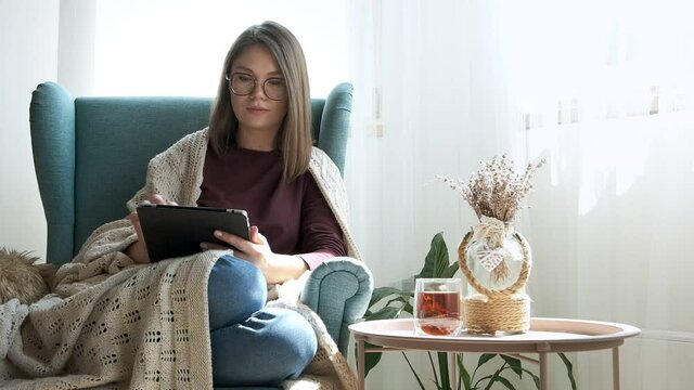 Young woman in eyeglasses using digital tablet, drinking tea, sitting in armchair at home