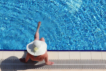 Woman in swimsuit and sun hat sitting on the edge of swimming pool and dangling her legs in water, top view. Summer vacation, tanning and leisure concept