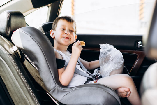 Child Sitting In A Car Seat And Eating Bread. Funny Little Boy With A Blurred Face Looking At The Camera.