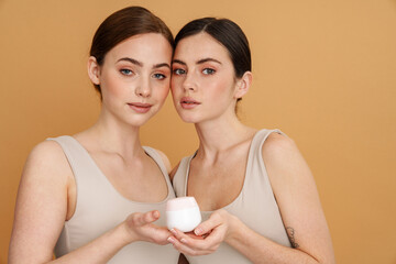 Young two women in undershirt showing face cream and looking at camera
