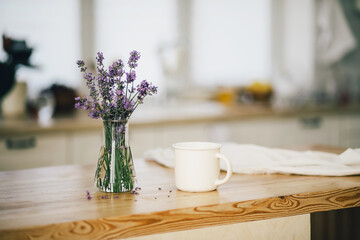 Vase with bunch of lavender and white cup of tea on a wooden table in a cozy kitchen.