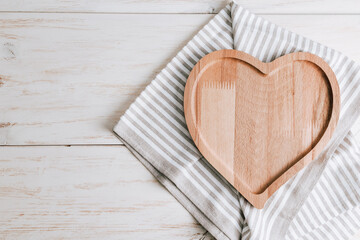 Heart on wooden background.
An empty heart-shaped plate with a kitchen napkin lie on the right on a wooden table with space for text on the left, close-up top view.