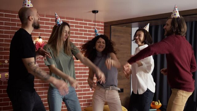Joyful friends in hats dancing on birthday party celebrating holiday at home, slowmo