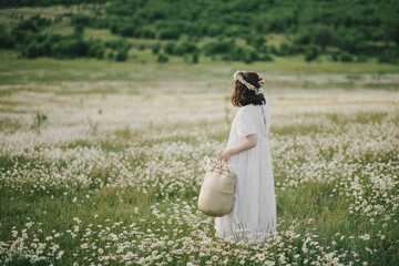 Young woman wearing white dress holding straw basket with flowers on chamomile field.