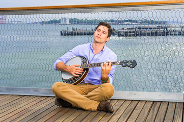 Against a fence, looking up, crossing legs  and thinking,  a young musician is sitting on the deck,...