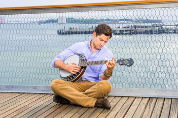 Lowering his head, crossing legs  and thinking,  a young musician is sitting on the deck, playing a banjo - obrazy, fototapety, plakaty