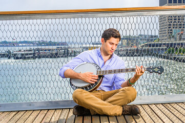 Lowering his head, crossing legs  and thinking,  a young musician is sitting on the deck, playing a...