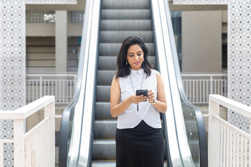 An Indian woman looking into her phone standing next to an escalator in an urban corporate setting