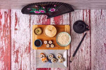 Top view image of maki, uramaki, black teapot, black chopsticks, california roll, dim sum steam basket, soy sauce, black fan and red and white table.
