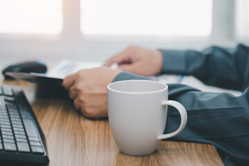 Close up white coffee cup and a man reading a newspaper on the work table in office