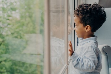 Lovely afro american kid in light blue shirt looking out of window