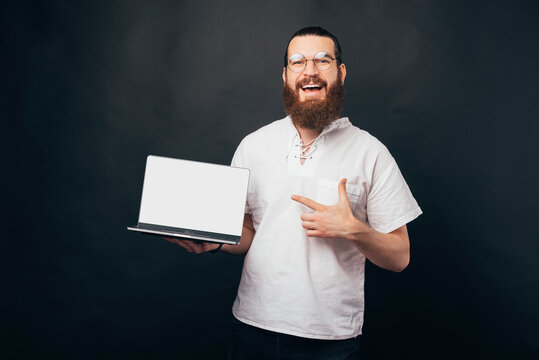 Excited man in glasses with beard pointing finger at blank screen laptop computer over dark background