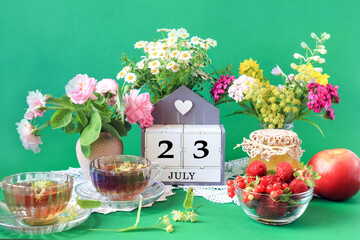  Calendar for July 23 : the name of the month of July in English, cubes with the number 23, bouquets of wild flowers, jam, fruit, cups of tea, white napkins, green background