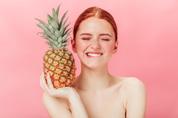 Front view of glad woman with pineapple posing with closed eyes. Studio shot of excited ginger girl holding fruit on pink background.