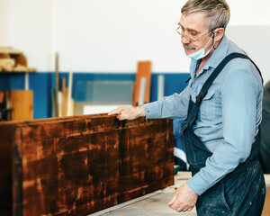 A gray-haired carpenter with glasses works with wood on a workbench in a carpentry shop. A...