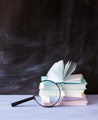 Stack of books and a magnifying glass on a wooden table against the background of a school board,...