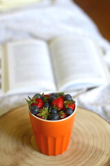 Cup of blueberries and strawberries and open book on a bed. Selective focus.