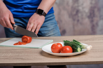 A man cuts a red tomato on a board. Cucumbers and tomatoes in a white plate