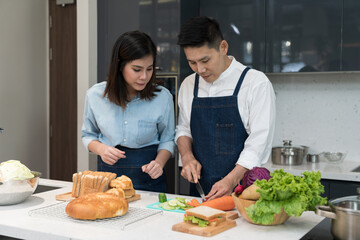 A happy man and young woman cut vegetables to cook healthy sandwiches on the kitchen island. Asian couple uses their free time to cooking food together on weekends in the kitchen. Culinary lifestyle.