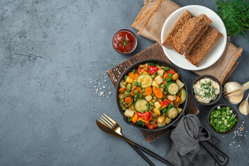 Lunch with vegetable stew, rye bread, sauces and herbs on a dark gray background.