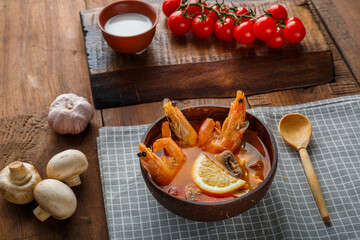 Tom yam soup with shrimps and coconut milk on the table on a checkered napkin next to vegetables and a wooden spoon.