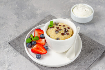 Mug cake with chocolate chips, fresh berries and cream on a gray concrete background. Copy space.