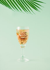 A wine glass with a drink and sea shells inside under a palm branch. Summer concept on pastel green backgrounds