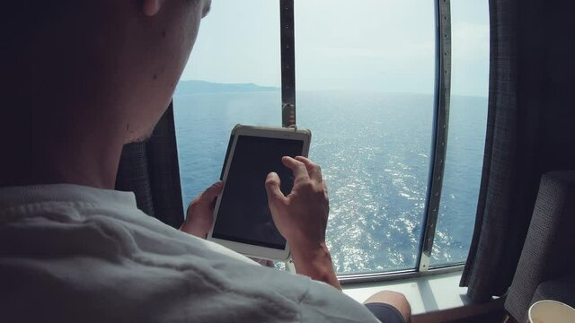 Male businessman clerk reading tablet computer on boat with modern technology while sailing on speedboat cruising ferry, view from cabin window