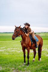 Fototapeta premium Young woman dressed in riding clothes and hat riding brown horse in green field