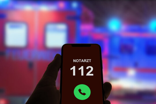 Person hält Smartphone in der Hand und ruft den Notarzt über die Notrufnummer 112, im Hintergrund Rettungswagen und Notarzt mit Blaulicht bei Nacht