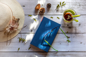 Top view of a book, glasses, glass of tea and hat on a worn white wooden table.