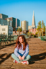 A young woman in jeans sits on a wooden floor on a pier in the morning with a view of downtown and...