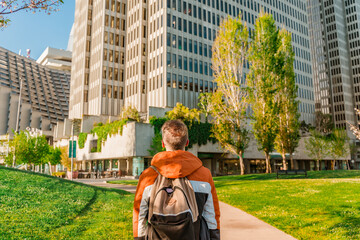 A young man with a backpack walks through downtown San Francisco with trees between business buildings