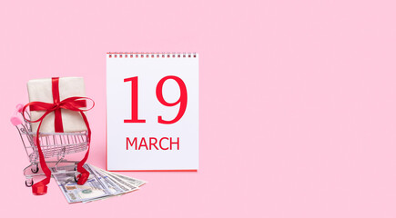 A gift box in a shopping trolley, dollars and a calendar with the date of 19 march on a pink background.